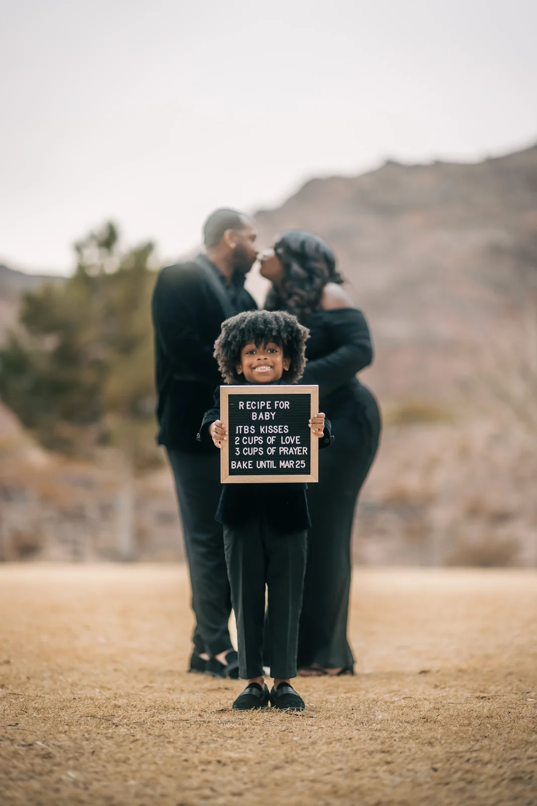 Child holding a baby announcement board while the parents kiss behind them during a maternity session in Henderson.