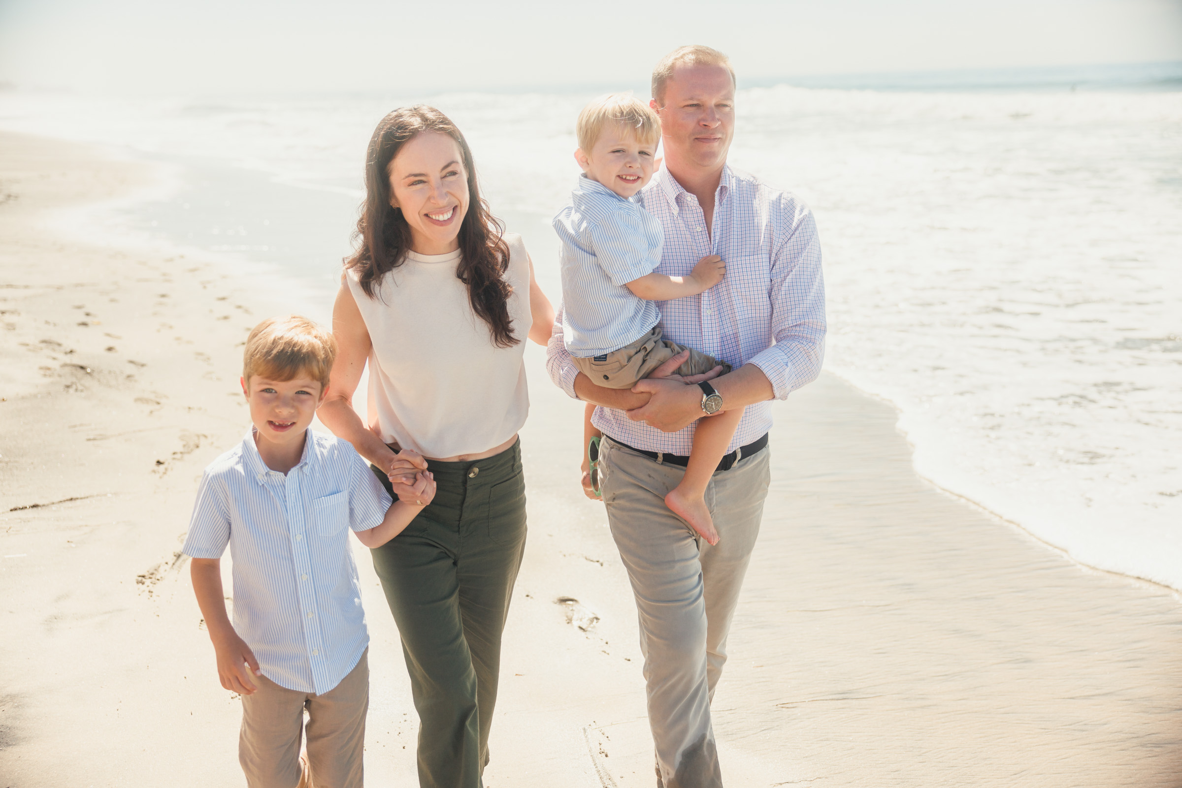 Parents and two boys walking along the shoreline, with the father carrying a toddler