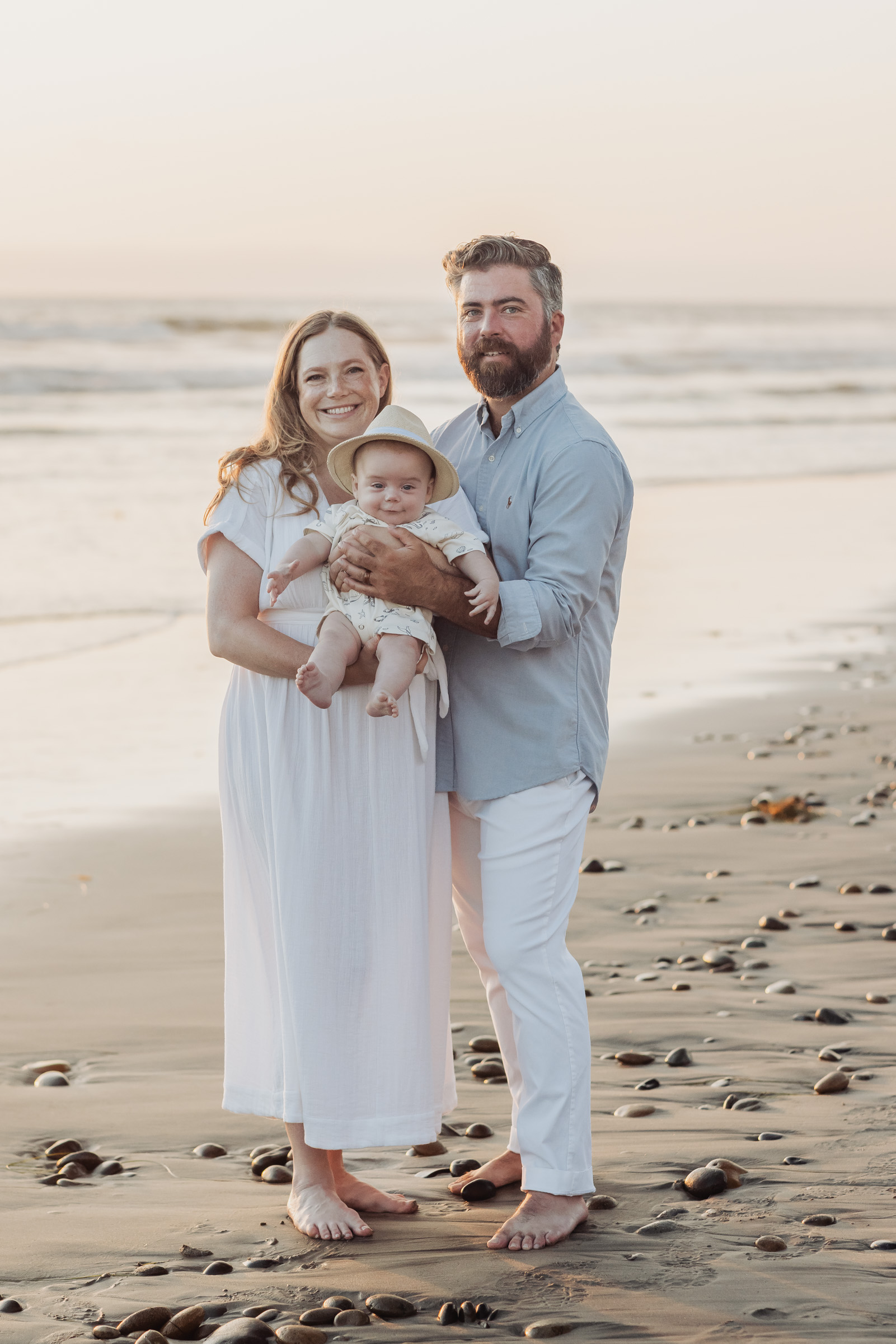 Parents holding their baby in a sun hat on a pebbly beach at sunset