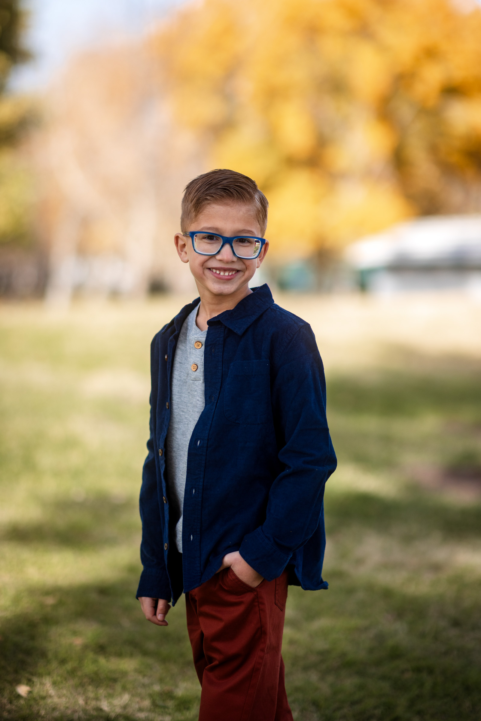 School-age boy with glasses in a navy shirt smiling with soft autumn foliage blurred behind him