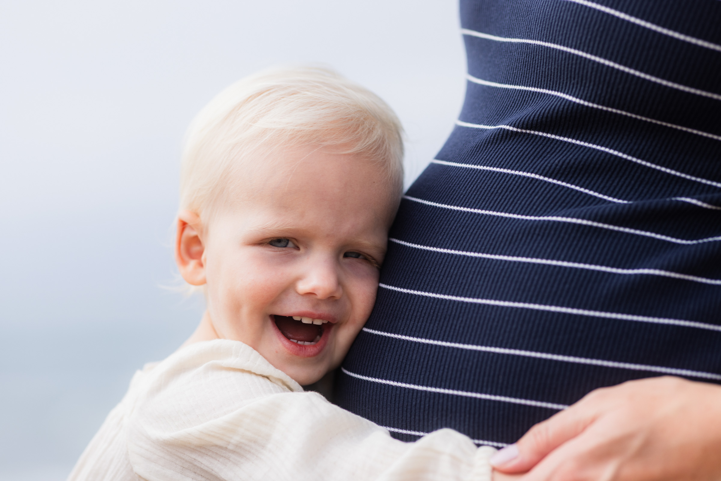 Toddler smiling with his cheek against a parent’s bump during a maternity session in Carlsbad.