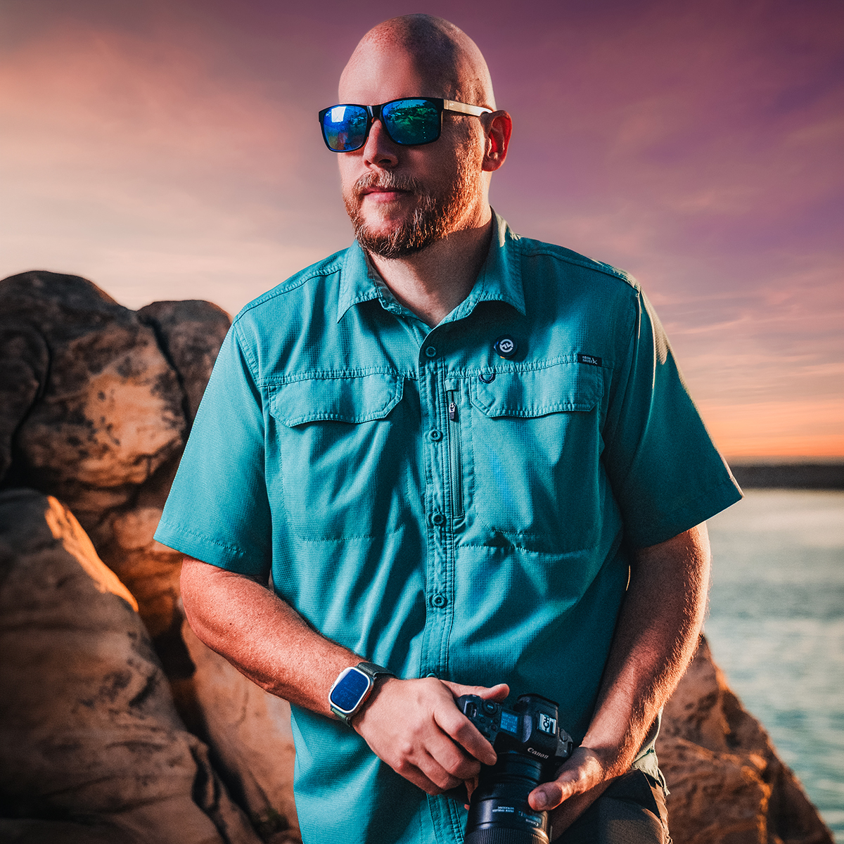 Joseph Blake holding a camera on the rocks at sunset by the water