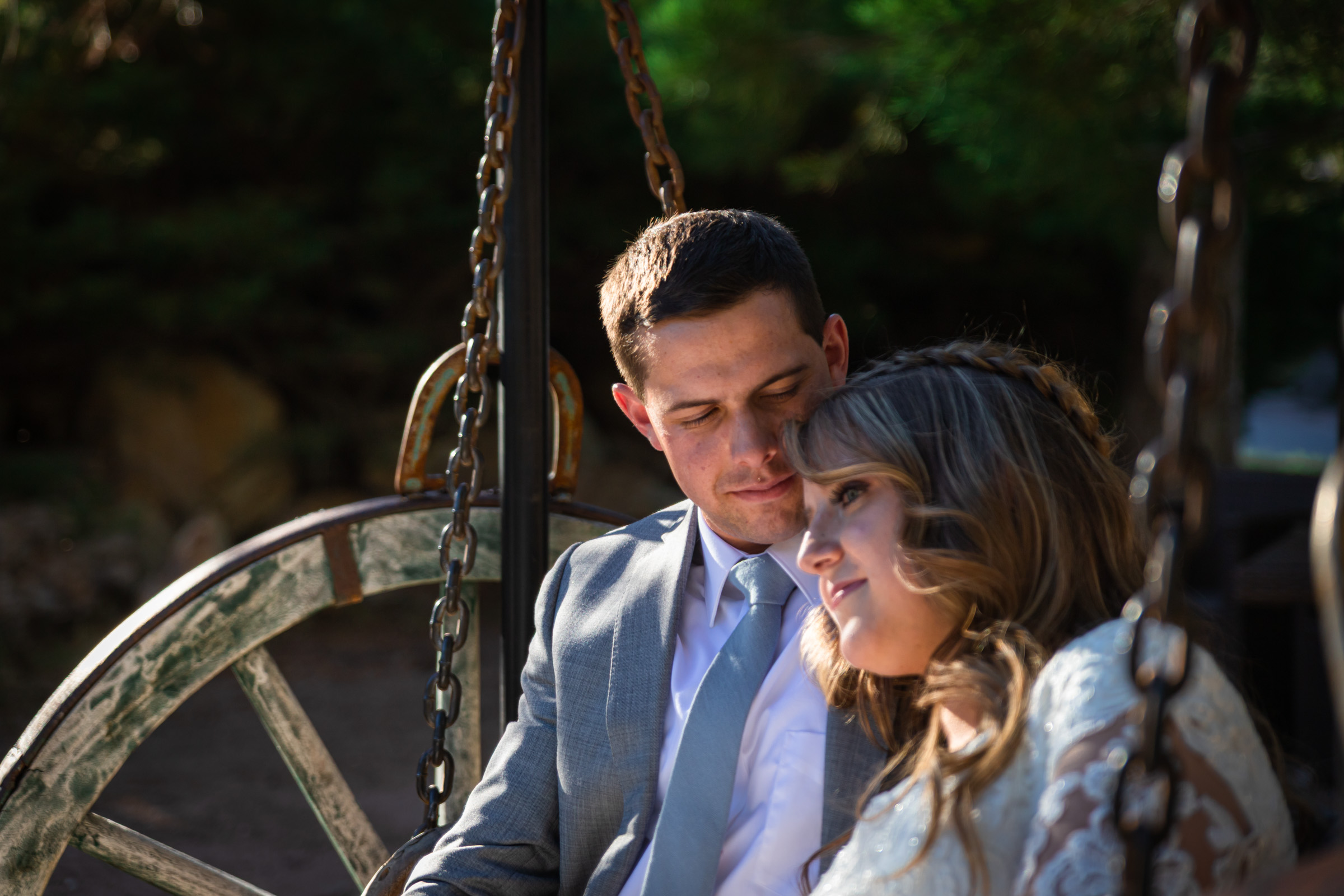 Bride and groom seated together on a rustic wagon-wheel swing in dappled forest light on Mount Charleston