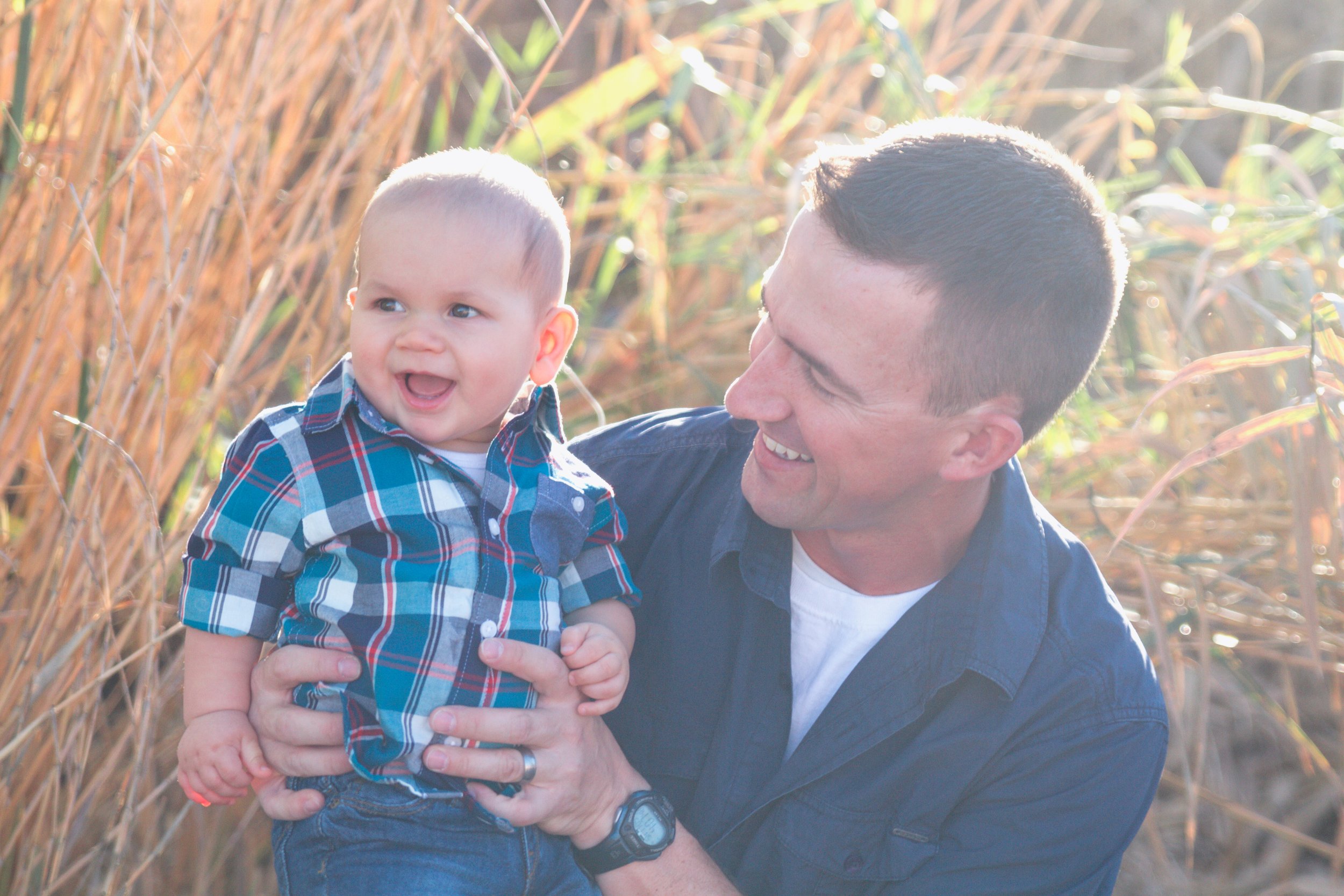 A smiling man holding a laughing baby in tall grass.