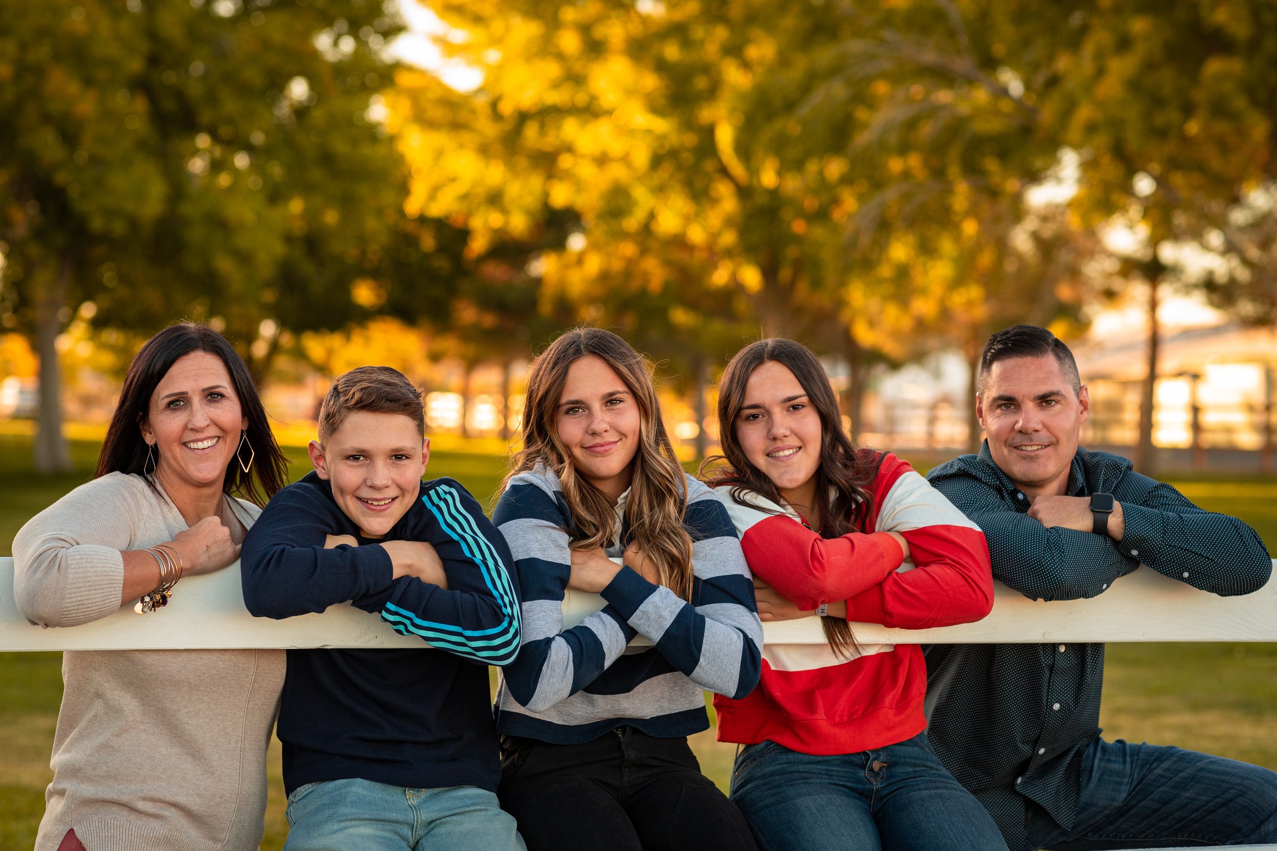 A family of five leaning on a white fence beneath autumn trees.