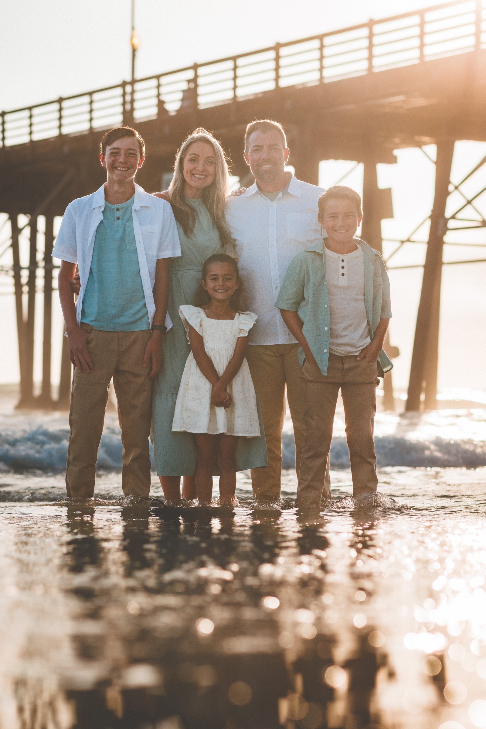 Family of five standing in the shallow surf in front of the Oceanside Pier at sunset
