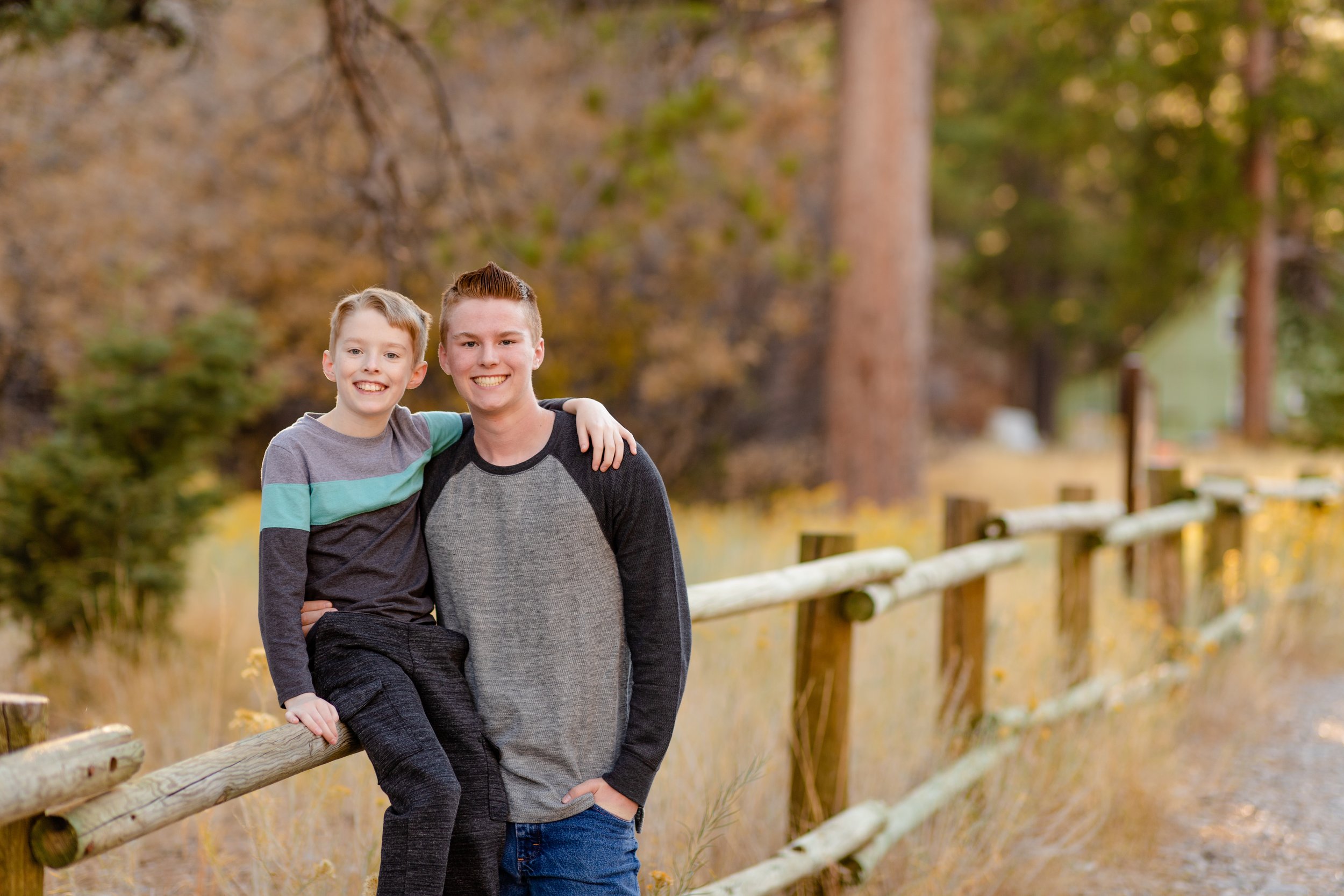 Two brothers smiling beside a wooden fence among autumn trees.