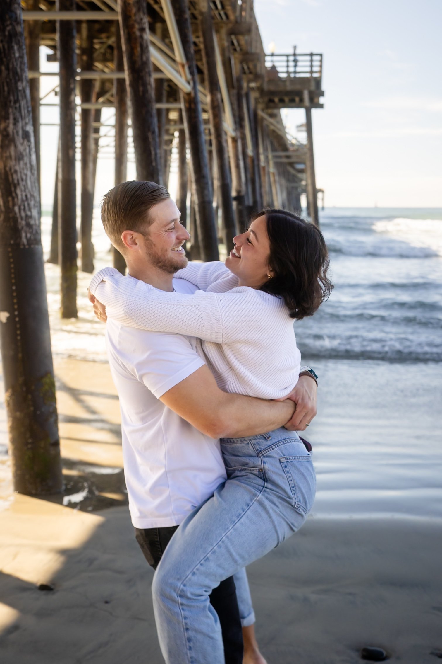 A man holding his partner in an embrace beneath a beach pier.